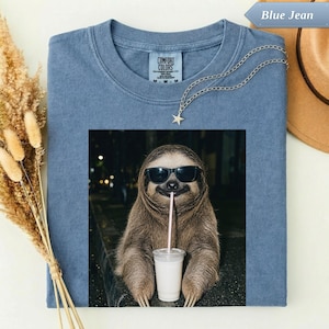May include: A blue jean coloured t-shirt featuring a graphic of a brown sloth wearing black sunglasses and holding a white cup with a straw. The shirt has a crew neckline and a tag that reads Comfort Colors. A silver star necklace is placed on top.