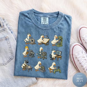 Puede incluir: Una camiseta azul con un estampado gráfico de nueve patos de dibujos animados realizando diversas tareas de ingeniería y mecánica, como soldar, dibujar y operar maquinaria. La camiseta tiene cuello redondo y una etiqueta de la marca Comfort Colors.