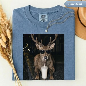 May include: A blue jean coloured t-shirt featuring a graphic of a brown deer wearing black sunglasses and drinking from a clear cup with a straw. The shirt has a Comfort Colors label at the neck and a silver star pendant necklace resting on the fabric.