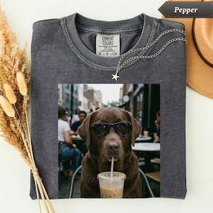 May include: A charcoal grey crew neck t-shirt featuring a graphic print of a brown Labrador dog wearing black sunglasses and drinking from a clear plastic cup with a straw. The shirt has a Comfort Colors label at the inner neckline.