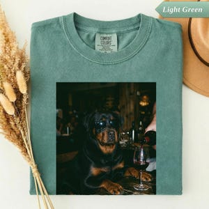Könnte beinhalten: Ein hellgrünes T-Shirt mit einem Grafikdruck eines schwarz-braunen Hundes, der eine schwarze Sonnenbrille trägt. Der Hund sitzt an einem Tisch mit einem Glas Rotwein. Das T-Shirt hat einen Rundhalsausschnitt und ein Comfort Colors Markenetikett.