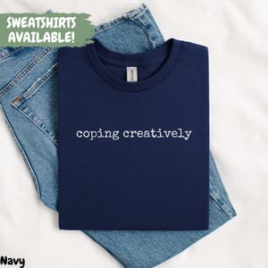 May include: Navy blue t-shirt with the phrase "coping creatively" in white, displayed on a pair of blue jeans. The text "SWEATSHIRTS AVAILABLE!" is in a green rectangle in the upper left corner. The word "Navy" is in the lower left corner.