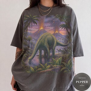 Może przedstawiać: Ciemnoszary t-shirt z krótkim rękawem z dużym nadrukiem graficznym przedstawiającym długoszyjego dinozaura pijącego z sadzawki w prehistorycznym krajobrazie z wybuchającymi wulkanami, palmami i mniejszymi dinozaurami pod fioletowo-pomarańczowym niebem.