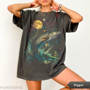 Peut inclure: Un t-shirt gris anthracite à manches courtes avec une impression graphique représentant un gros poisson sautant hors de l'eau devant une pleine lune jaune et des éclairs. Le t-shirt a un col rond et une coupe ample et décontractée.
