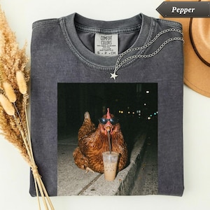 Puede incluir: Camiseta Comfort Colors gris oscuro con un gráfico de un pollo con gafas de sol bebiendo de una taza. Un collar plateado con un colgante de estrella está sobre la camiseta. La palabra "Pepper" está en una flecha negra.