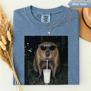 Könnte beinhalten: Ein blaues T-Shirt mit einer Grafik eines braunen Bibers, der eine schwarze Sonnenbrille trägt und aus einem durchsichtigen Plastikbecher mit Strohhalm trinkt. Das T-Shirt hat einen Rundhalsausschnitt und ein Comfort Colors Markenetikett.