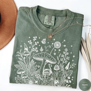 May include: An olive green t-shirt featuring a white line art graphic of mushrooms, wildflowers, and three gnomes. The shirt has a Comfort Colors brand label at the inner neckline and is displayed with a gold chain necklace and a brown hat.