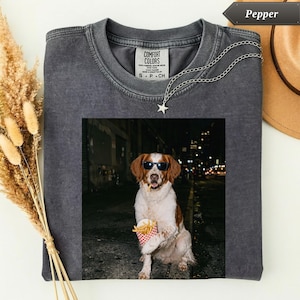 Puede incluir: Una camiseta de cuello redondo gris carbón con un estampado gráfico de un perro marrón y blanco que lleva gafas de sol negras y sostiene un pequeño recipiente rojo y blanco de patatas fritas en sus patas sobre una calle oscura.