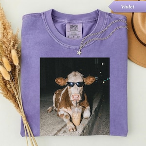 Purple t-shirt with a cow wearing sunglasses drinking a milkshake.