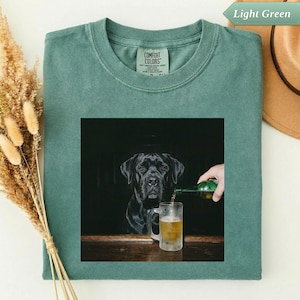 Könnte beinhalten: Hellgrünes Comfort Colors T-Shirt mit einem quadratischen Aufdruck eines schwarzen Hundes mit einem Bierkrug. Eine Hand gießt Bier in den Krug. Das Shirt hat einen Rundhalsausschnitt und kurze Ärmel. Ein Hut ist im Hintergrund zu sehen.