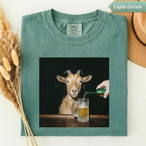 Puede incluir: Camiseta verde claro Comfort Colors con una imagen cuadrada de una cabra y una jarra de cerveza que se está llenando. La cabra está mirando al espectador. La camiseta es de color verde claro liso.