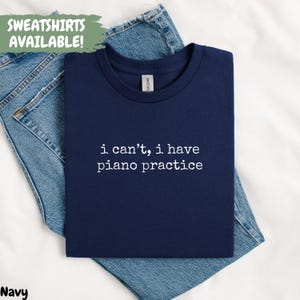 May include: Navy blue t-shirt with the text "i can't, i have piano practice" in white. The shirt is displayed on a pair of blue jeans. The words "SWEATSHIRTS AVAILABLE!" are in a green box in the upper left corner.