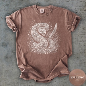 Może przedstawiać: Brązowy t-shirt z krótkim rękawem z białą grafiką przedstawiającą zwiniętego grzechotnika z wysuniętym językiem, otoczonego małymi okrągłymi kropelkami. Koszulka ma okrągły dekolt i metkę marki Comfort Colors przy wewnętrznym kołnierzyku.