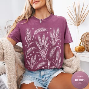 Könnte beinhalten: Ein beerenfarbenes Kurzarm-T-Shirt mit einem weißen Grafikdruck aus verschiedenen Weizenähren und kleinen Sternen. Das T-Shirt hat einen Rundhalsausschnitt und eine lockere Passform, die eine detaillierte botanische Getreideillustration zeigt.