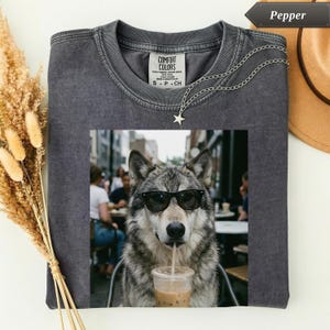 May include: A charcoal gray crew neck t-shirt featuring a graphic print of a wolf wearing black sunglasses and drinking from a clear plastic cup with a straw. The shirt has a Comfort Colors label at the inner neckline and a silver star necklace.