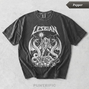 May include: A charcoal gray short-sleeve t-shirt featuring a white graphic print. The design displays the word LESBIAN in a metal-style font above a warrior figure holding a spear, flanked by two winged serpents, with lightning and skulls below.