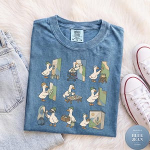 May include: A blue short-sleeve t-shirt featuring a graphic print of nine white ducks performing various painting and artistic tasks. The shirt has a crew neckline and a Comfort Colors brand label printed on the inside of the collar.