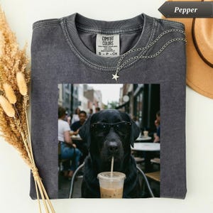 May include: A charcoal grey crew neck t-shirt featuring a graphic print of a black Labrador dog wearing black sunglasses and drinking from a clear plastic cup with a straw. The shirt has a Comfort Colors label at the inner neckline.