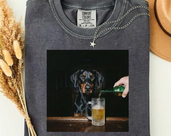Puede incluir: Una camiseta Comfort Colors gris oscuro con un gráfico de un Doberman Pinscher y una jarra de cerveza. El perro mira al espectador mientras se vierte cerveza en la jarra. Un collar plateado con un colgante de estrella está sobre la camisa.