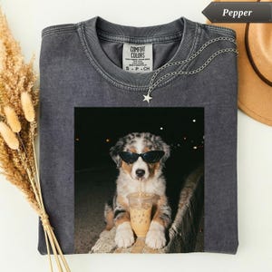 May include: Dark gray Comfort Colors t-shirt featuring a graphic of a dog wearing sunglasses and drinking an iced coffee. A silver necklace with a star pendant hangs on the shirt. The shirt has a crew neck and short sleeves.