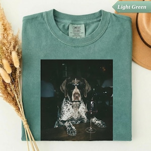 May include: A light green t-shirt featuring a graphic print of a spotted dog wearing black sunglasses, sitting at a table with a wine glass. The shirt has a Comfort Colors brand label at the inner neckline and a crew neck collar design.