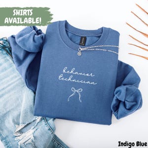 May include: Indigo blue sweatshirt with the words "behavior technician" in white script and a bow graphic. The text "SHIRTS AVAILABLE!" is in a green box. A silver necklace is draped across the top of the sweatshirt.