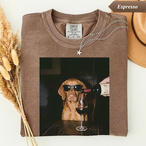 Op de afbeelding: Een bruin t-shirt met ronde hals en een grafische print van een hond met een zwarte zonnebril terwijl er rode wijn in een glas wordt geschonken. Het t-shirt heeft een Comfort Colors-label aan de binnenkant van de halslijn.