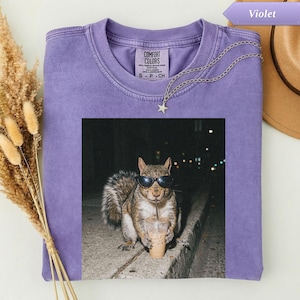 May include: A violet Comfort Colors t-shirt featuring a graphic of a squirrel wearing sunglasses and drinking an iced coffee. A silver necklace with a star pendant is draped on the shirt. The shirt has a crew neck and short sleeves.