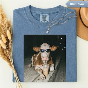 Blue t-shirt with a cow wearing sunglasses drinking from a cup.