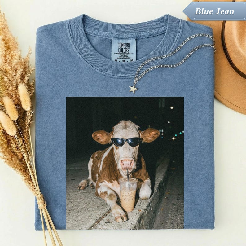 Blue t-shirt with a cow wearing sunglasses drinking from a cup.