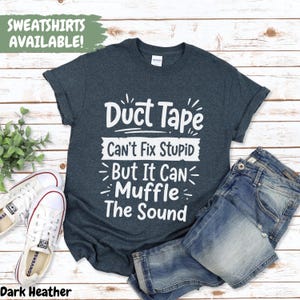 May include: Dark heather t-shirt with the text "Duct Tape Can't Fix Stupid But It Can Muffle The Sound" in white. The shirt is paired with blue jeans and white sneakers. The words "SWEATSHIRTS AVAILABLE!" are in a green box.