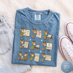 Könnte beinhalten: Ein blaues T-Shirt mit einem Grafikdruck von neun Cartoon-Hirschfiguren, die verschiedene Büro- und Designaufgaben ausführen, darunter Präsentationen auf Whiteboards, die Arbeit an Laptops und das Überprüfen von Dokumenten. Das Shirt hat ein Comfort Colors Etikett.