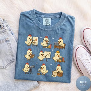 May include: A blue short-sleeved t-shirt featuring a graphic print of nine yellow chicks performing various professional tasks, including using a laptop, a telescope, and a whiteboard. The shirt has a Comfort Colors brand label at the neck.