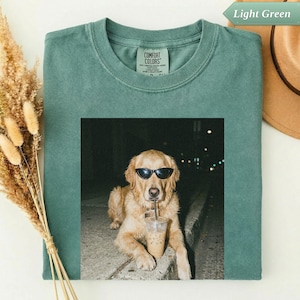 Op de afbeelding: Lichtgroen Comfort Colors T-shirt met een foto van een golden retriever met een zonnebril op en een ijskoffie drinkend. De hond zit 's nachts op een betonnen stoeprand. Een hoed en droogbloemen staan op de achtergrond.