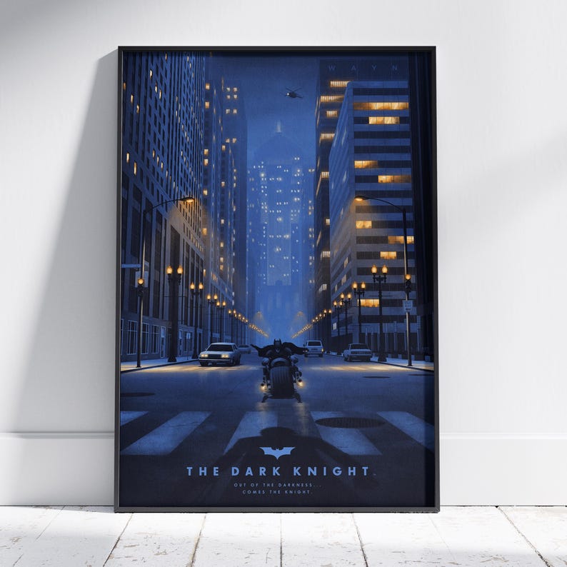 May include: A framed vertical poster depicting a dark city street at night with tall buildings and glowing windows. A figure on a motorcycle rides down the centre of the road. The text reads THE DARK KNIGHT and OUT OF THE DARKNESS COMES THE KNIGHT.