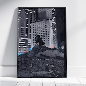 May include: A framed vertical poster featuring a dark, monochromatic cityscape with a figure in a cape standing atop a pile of rubble. The text THE DARK KNIGHT is printed in the centre, with smaller text below and a bat symbol icon.