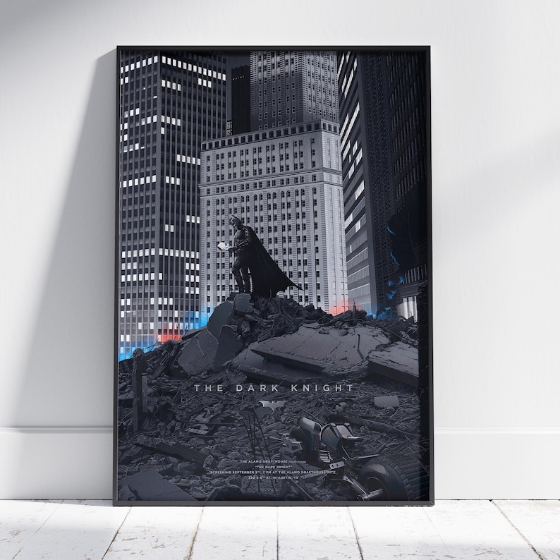 May include: A framed vertical poster featuring a dark, monochromatic cityscape with a figure in a cape standing atop a pile of rubble. The text THE DARK KNIGHT is printed in the centre, with smaller text below and a bat symbol icon.