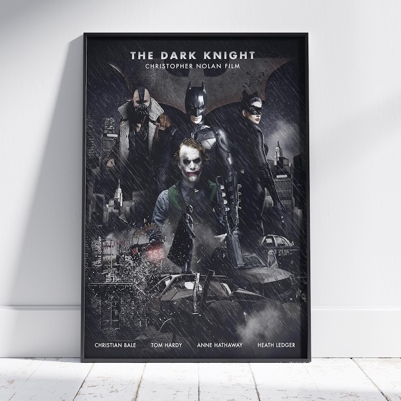 May include: A framed movie poster for The Dark Knight, a Christopher Nolan film. The artwork features Batman, Bane, Catwoman, and the Joker in a dark, rainy city setting with a bat symbol in the background. Cast names are printed at the bottom.