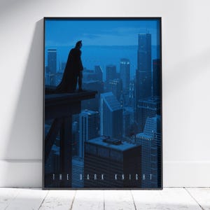 May include: A framed vertical poster featuring a dark blue cityscape at night with a silhouette of a caped figure standing on a ledge overlooking the buildings. The text THE DARK KNIGHT is printed in white capital letters at the bottom of the image.