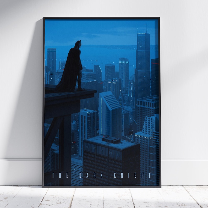 May include: A framed vertical poster featuring a dark blue cityscape at night with a silhouette of a caped figure standing on a ledge overlooking the buildings. The text THE DARK KNIGHT is printed in white capital letters at the bottom of the image.