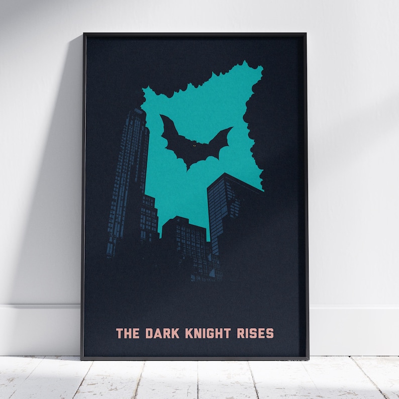 May include: A rectangular framed poster featuring a dark blue cityscape silhouette against a bright teal background. A black bat symbol is centred in the teal area. The text THE DARK KNIGHT RISES is printed in light pink at the bottom of the poster.