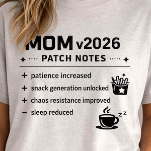 May include: A light gray t-shirt with the text "MOM v2026 PATCH NOTES" and a list of humorous updates: patience increased, snack generation unlocked, chaos resistance improved, and sleep reduced. The shirt features a coffee cup and french fries graphic.