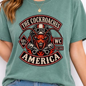 May include: A sage green short-sleeve t-shirt featuring a graphic of a red insect wearing goggles, flanked by crossed pistons and flames. The text reads THE COCKROACHES, 69% BUG, WC, EST. 2026, and AMERICA in a bold, vintage-inspired banner design.