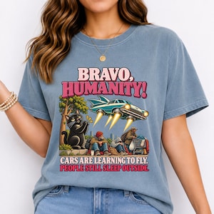 May include: A blue t-shirt featuring a graphic print with the text BRAVO, HUMANITY! CARS ARE LEARNING TO FLY. PEOPLE STILL SLEEP OUTSIDE. The design shows a flying car, a black cat, and people sitting on the ground with a shopping cart.