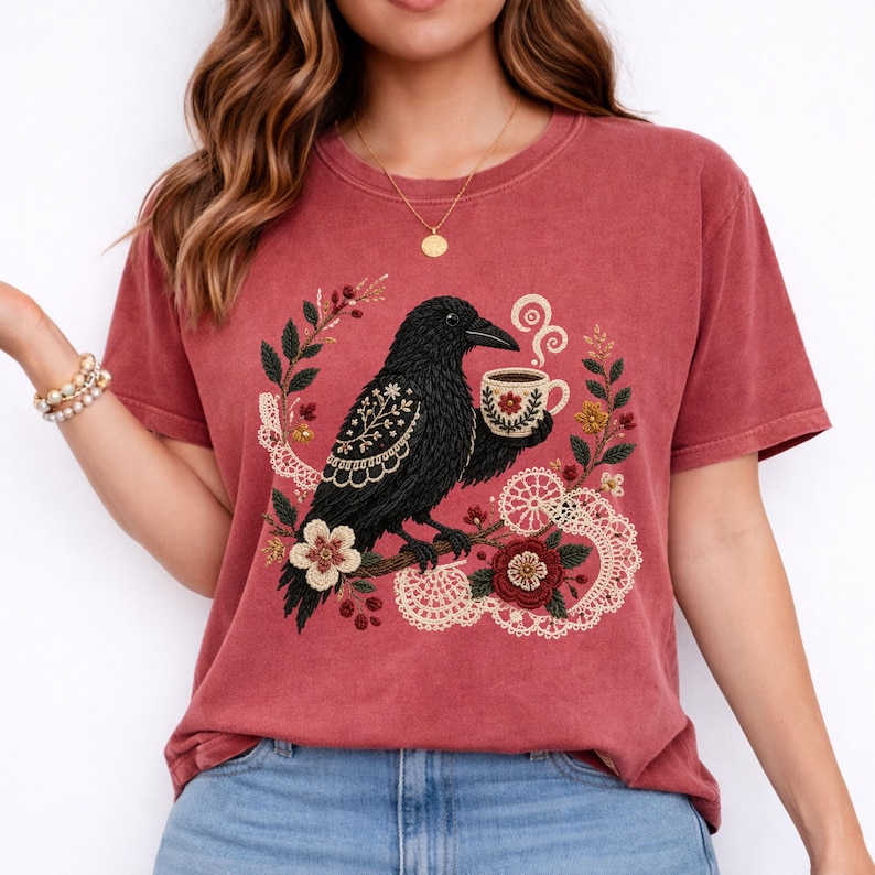 Black crow holding a coffee cup with floral and lace details on a crimson Comfort Colors t-shirt