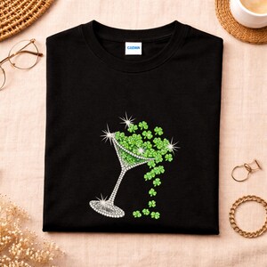Black t-shirt featuring a sparkling martini glass design filled with green shamrocks. The glass is embellished with rhinestones, and the shamrocks cascade down the shirt. The shirt has a classic crew neck and short sleeves.