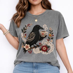 Gray t-shirt featuring a black raven holding a coffee cup, surrounded by floral and lace embroidery. The design includes red, white, and yellow accents. The shirt is a comfortable, casual style.