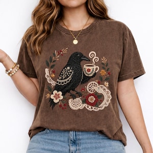 Black crow holding a coffee cup with floral and lace details on a brown Comfort Colors t-shirt