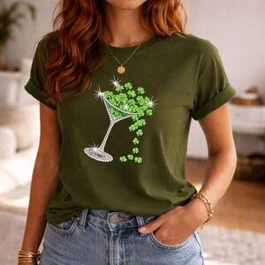 Olive green t-shirt featuring a rhinestone martini glass filled with green shamrocks. The glass design has a sparkling rim and stem. The shamrocks are spilling out of the glass. The shirt is a casual style.