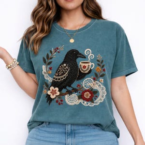Teal-colored t-shirt featuring an embroidered black crow holding a coffee cup. The crow is perched on a branch surrounded by floral and lace details. The design includes red, white, and yellow flowers, and the shirt has a relaxed fit.